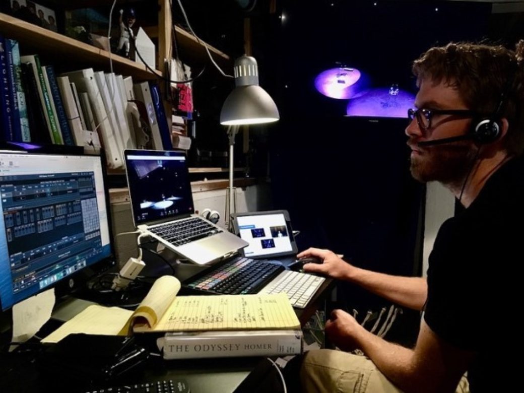 A figure wearing headphones works on multiple computer monitors with a cluttered desk