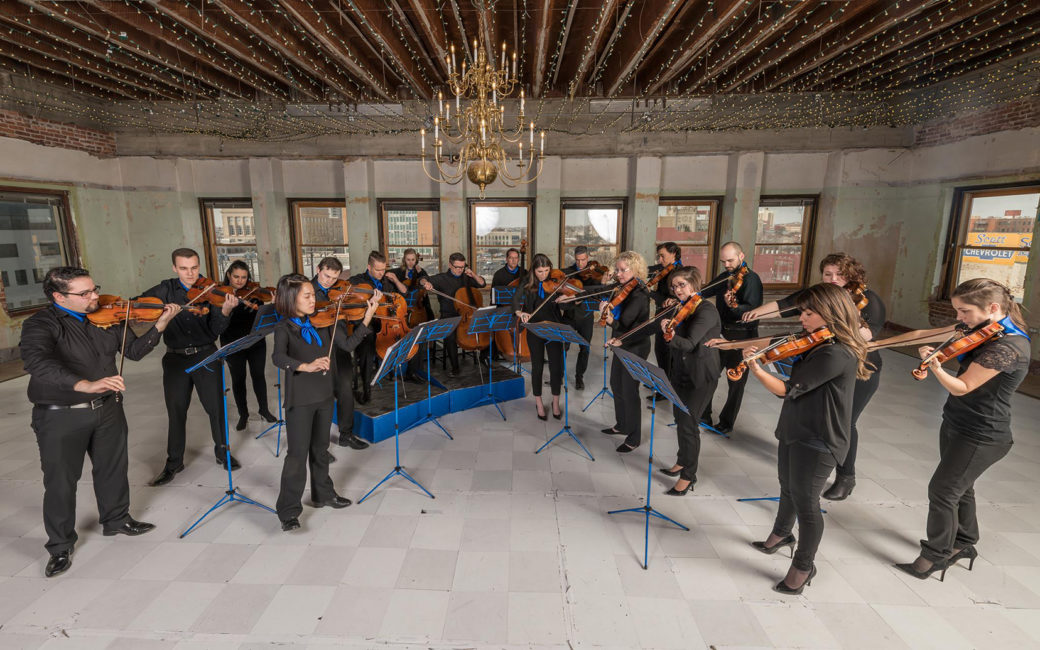 Orchestra members playing while standing in a concrete room with a chandelier above.