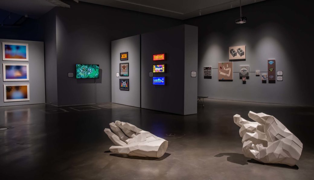 Two white, geometric hand sculptures lay on a concrete floor in a gallery with gray walls featuring screens and artworks while figures look on