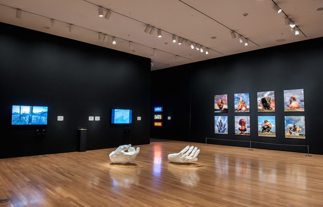 A pair of white, sculptural hands lay on a wood floor in a gallery with black walls featuring screens and artworks