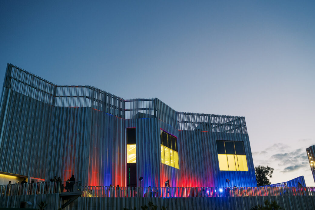 An angular, metal building with large windows is illuminated by red and blue lights