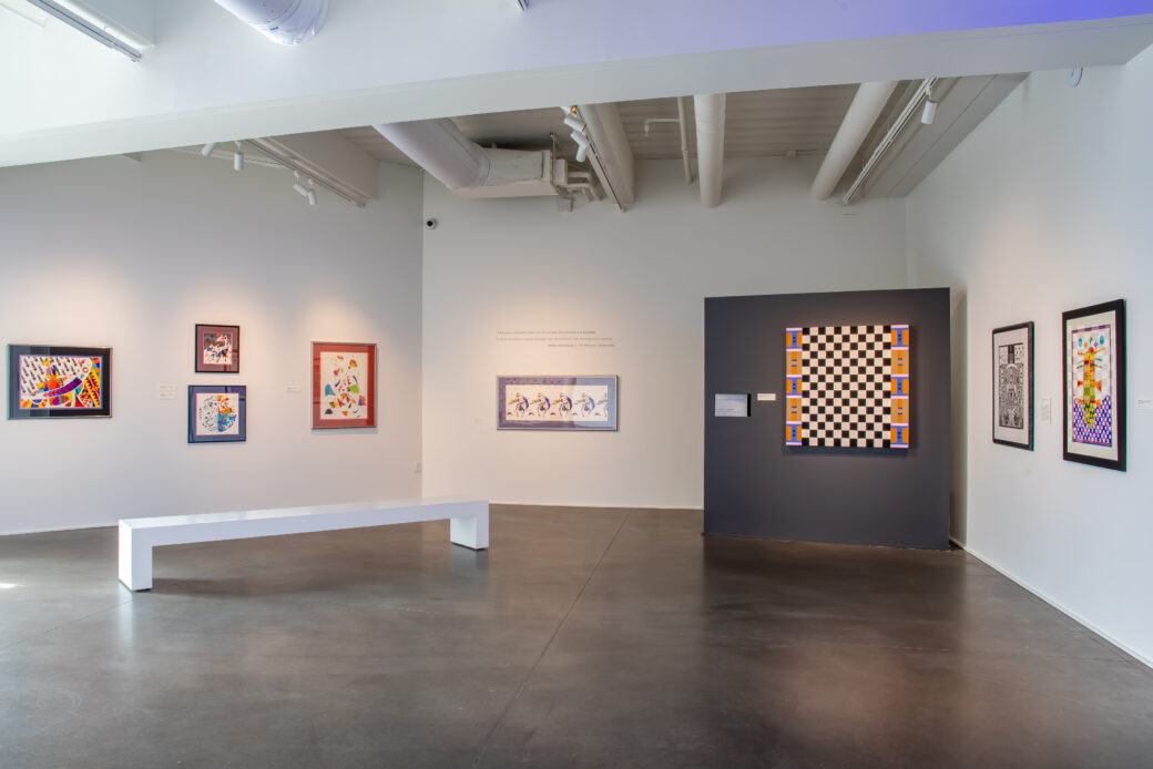 Multiple brightly colored abstract and geometrical paintings and drawings hang in a gallery