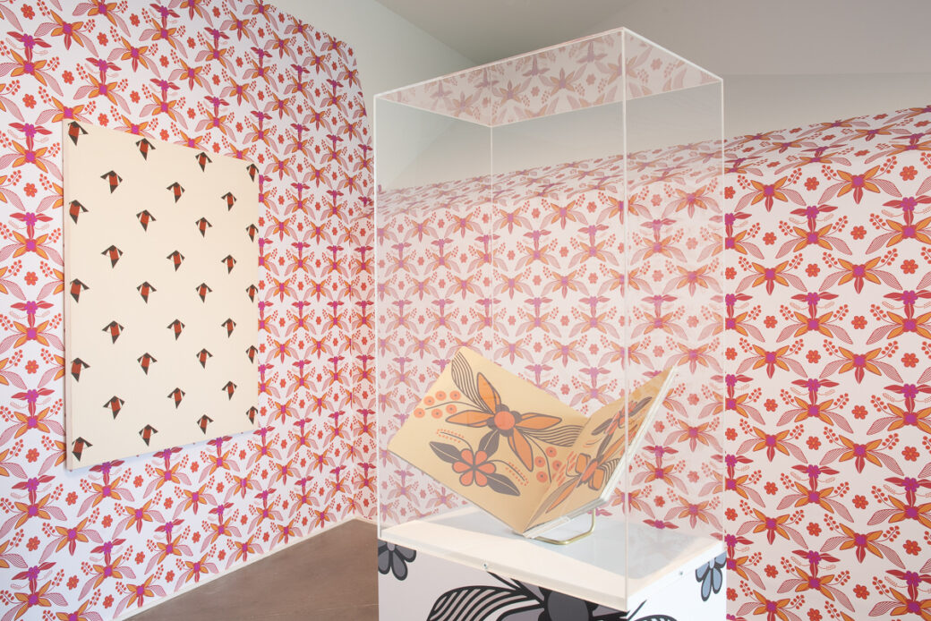 A book printed with floral patterns is displayed on a plinth. Walls covered in pink and orange florals with a painting of a repeating pattern hanging on the left are in the background.