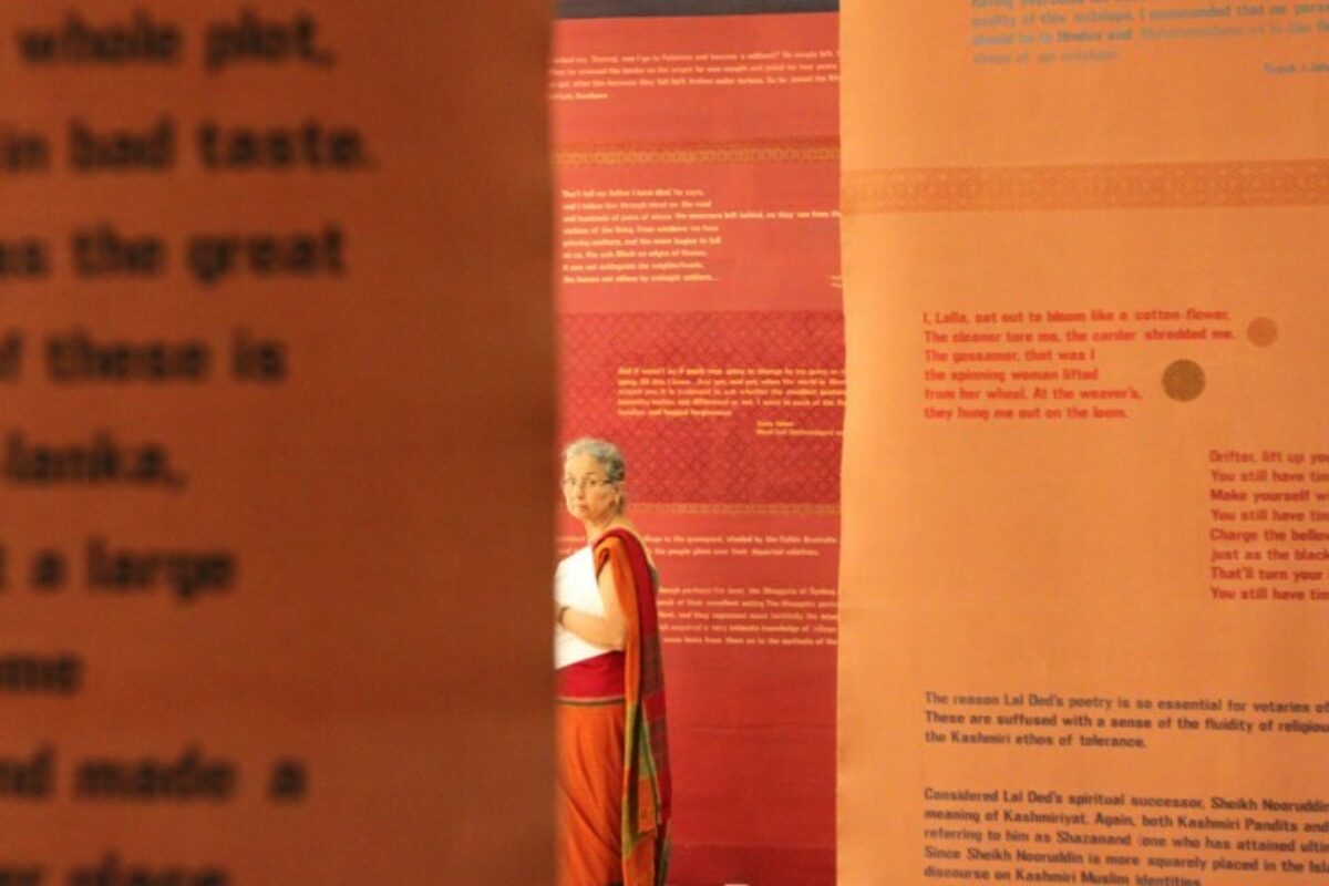 An older woman in an orange draped garment walks between walls printed with text