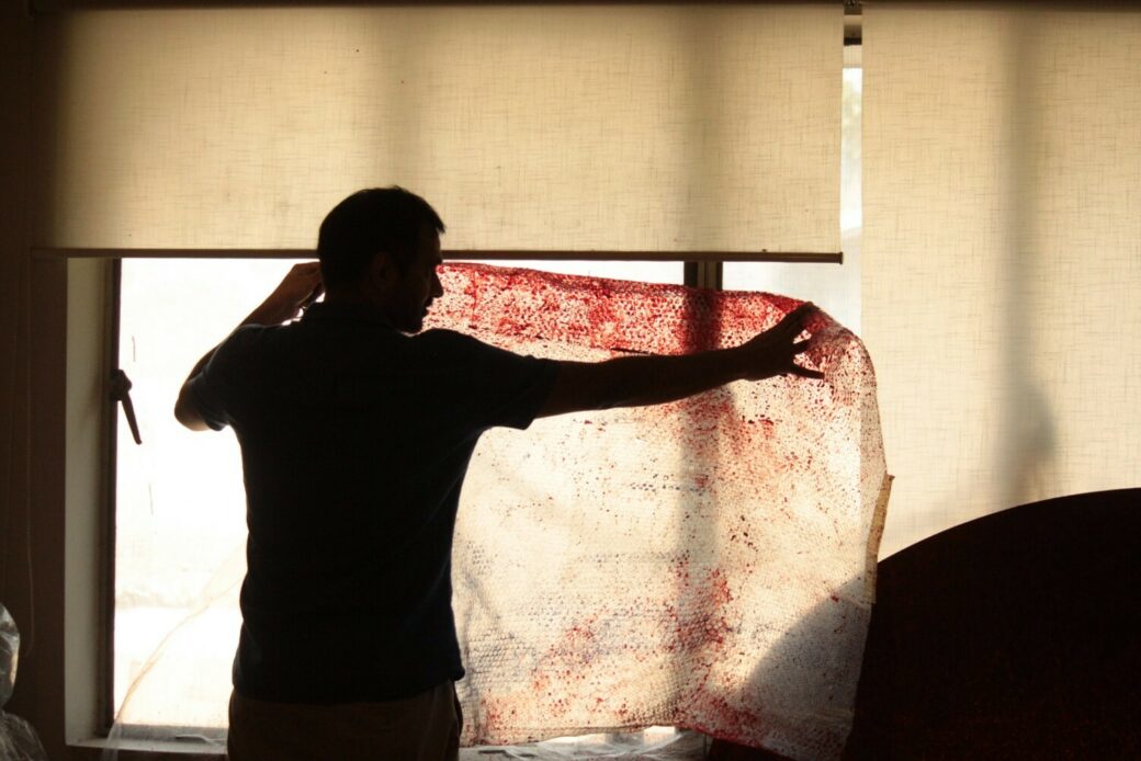 A man's silhouette is visible against a lit window as he works with a large sheet of paper