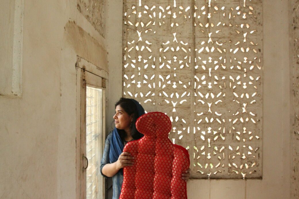 A woman holds a life-size red pillow in the shape of a person while standing against a fretwork wall