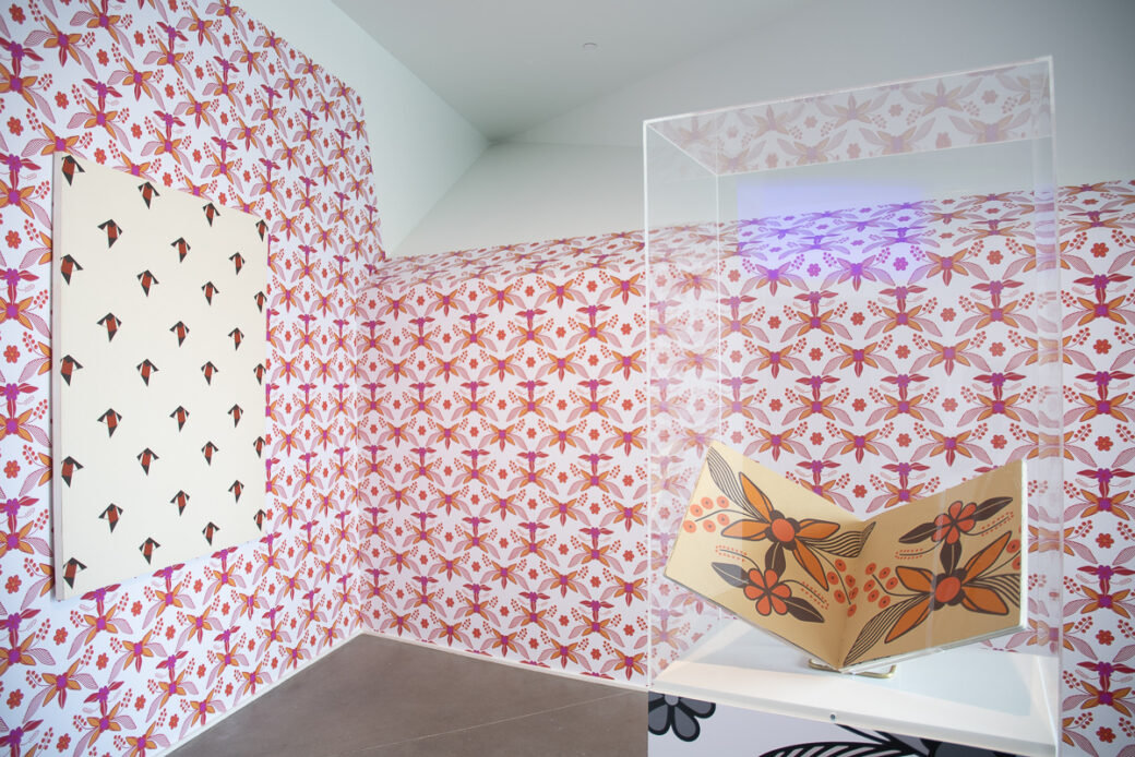 A canvas covered in a repeating pattern hangs on a wall plastered in brightly patterned wallpaper. A book featuring large scale patterns is displayed on a plinth.