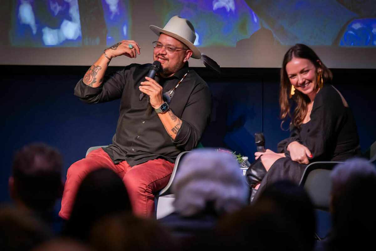 A man in red pants, a black shirt and hat with a feather talks into a microphone while a woman sitting next to him laughs