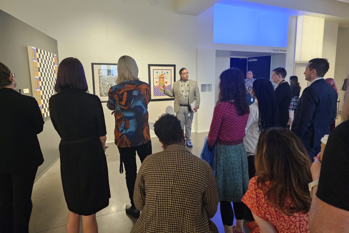 A group of people stand in a gallery while a man in a suit points to an artwork