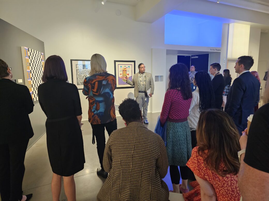 A group of people stand in a gallery while a man in a suit points to an artwork