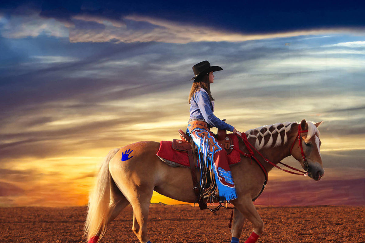 A photograph in which a cowgirl in ornamented chaps rides a chestnut horse with a white mane across a colorful skyline