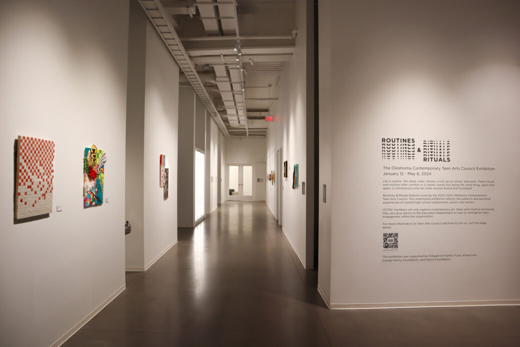 A hallway of an art gallery with white walls has several pieces hanging in multimedia and varrying colors. A title wall to the right reads ROUTINES & RITUALS in a pattern-like logo stacked on top of itself.