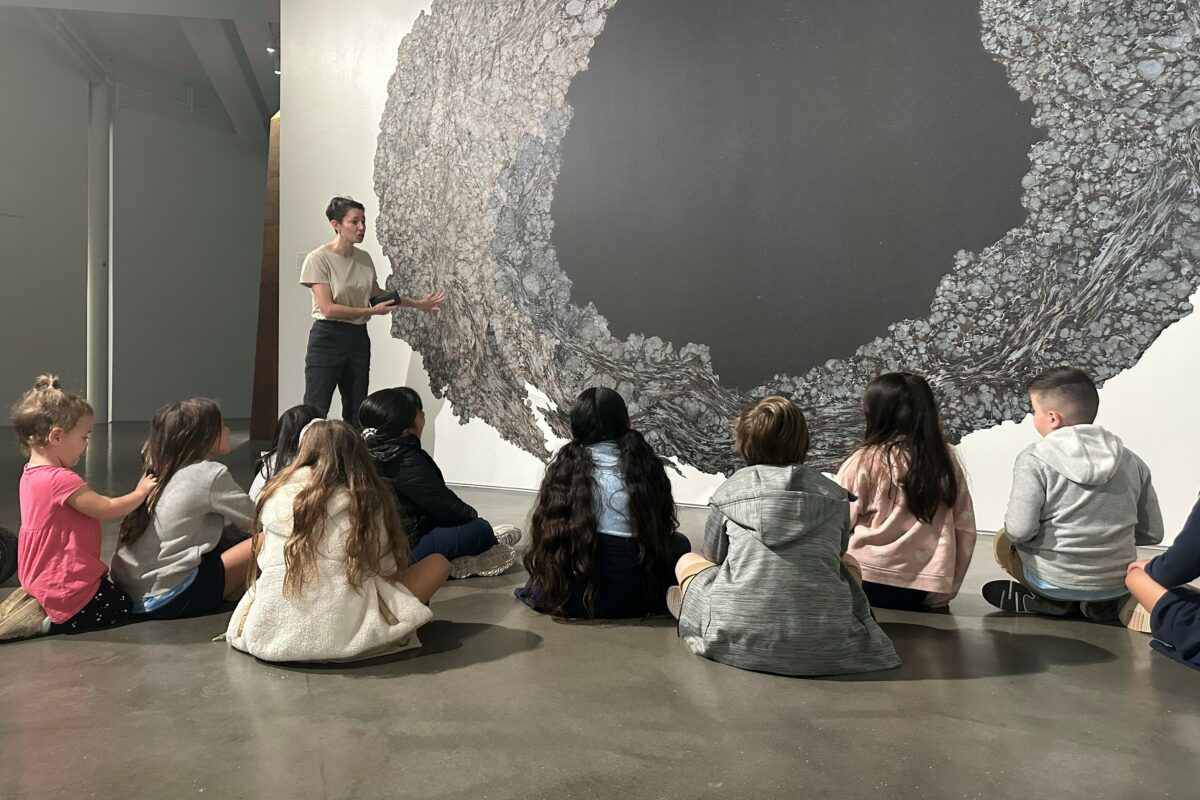 A group of children sit on the floor in front of a portal-like mural with an adult standing next to it