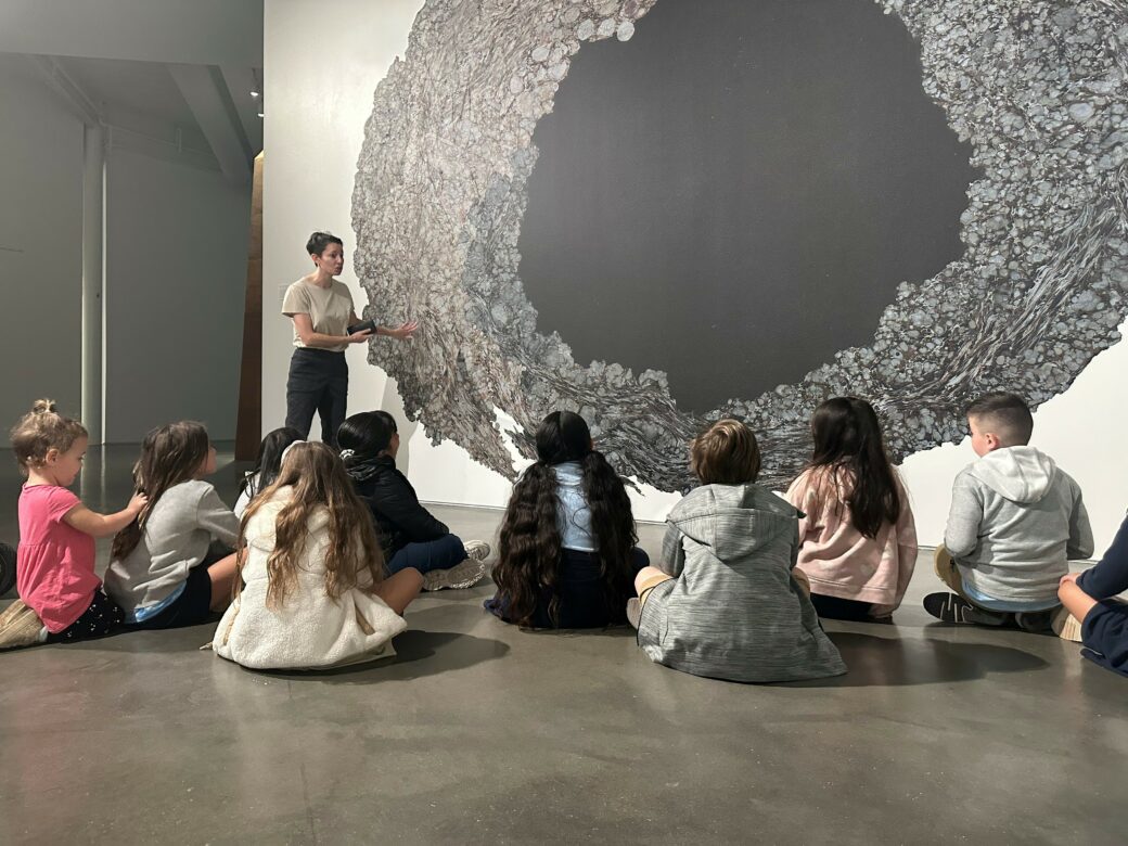 A group of children sit on the floor in front of a portal-like mural with an adult standing next to it