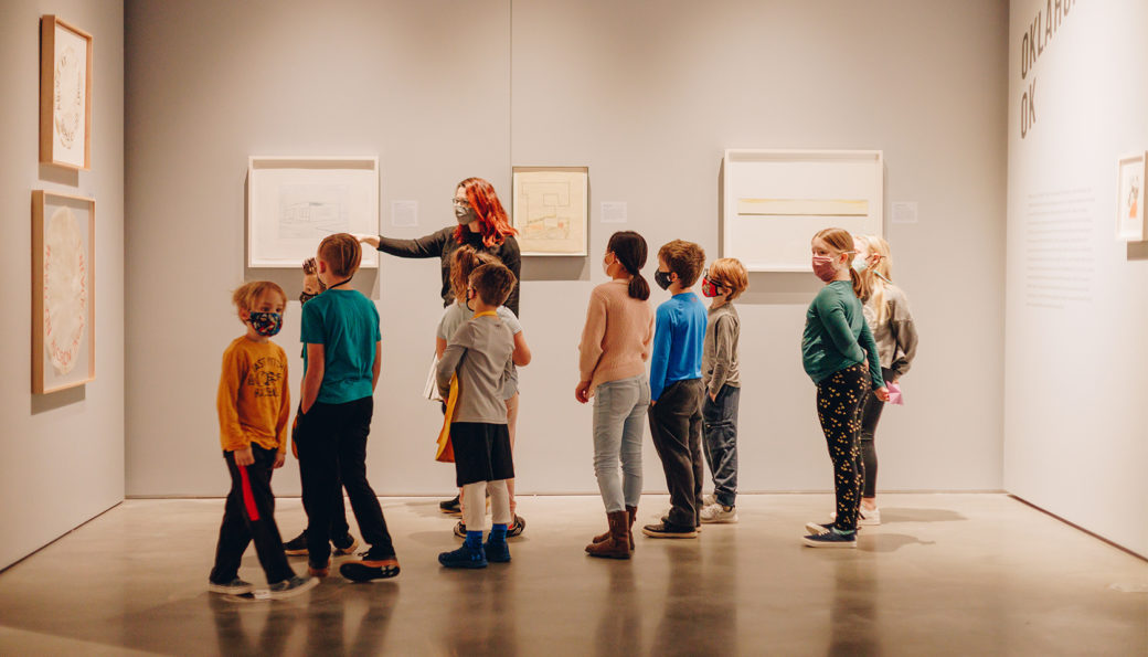An adult guides a group of masked children through a gray-walled gallery with concrete floors