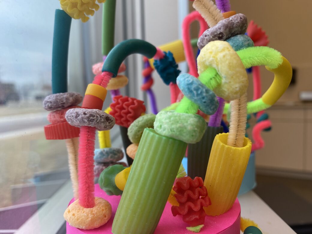 Dried pasta dyed green, blue, yellow and pink are combined with colorful Fruit Loops, wrapped and hanging from pipe cleaners to form a pasta and cereal sculpture.