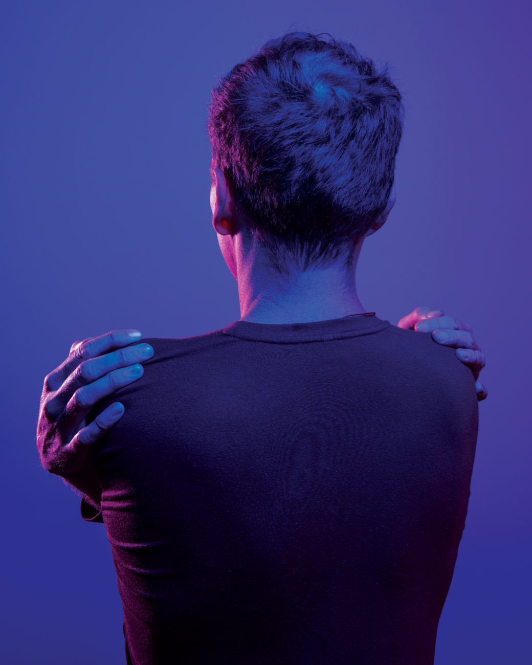 A person with their hands crossed, grabs their shoulders, facing away from the viewer (image in hues of blue)