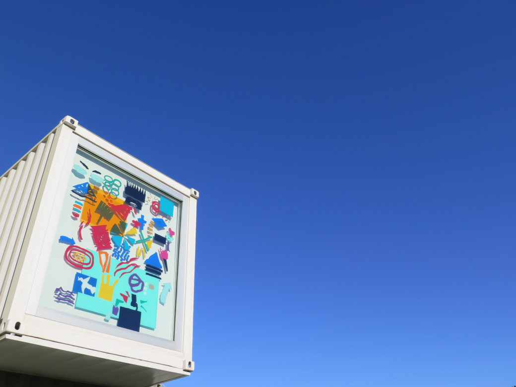 A window of a white shipping container building features a multicolored, hieroglyphic mural in vinyl