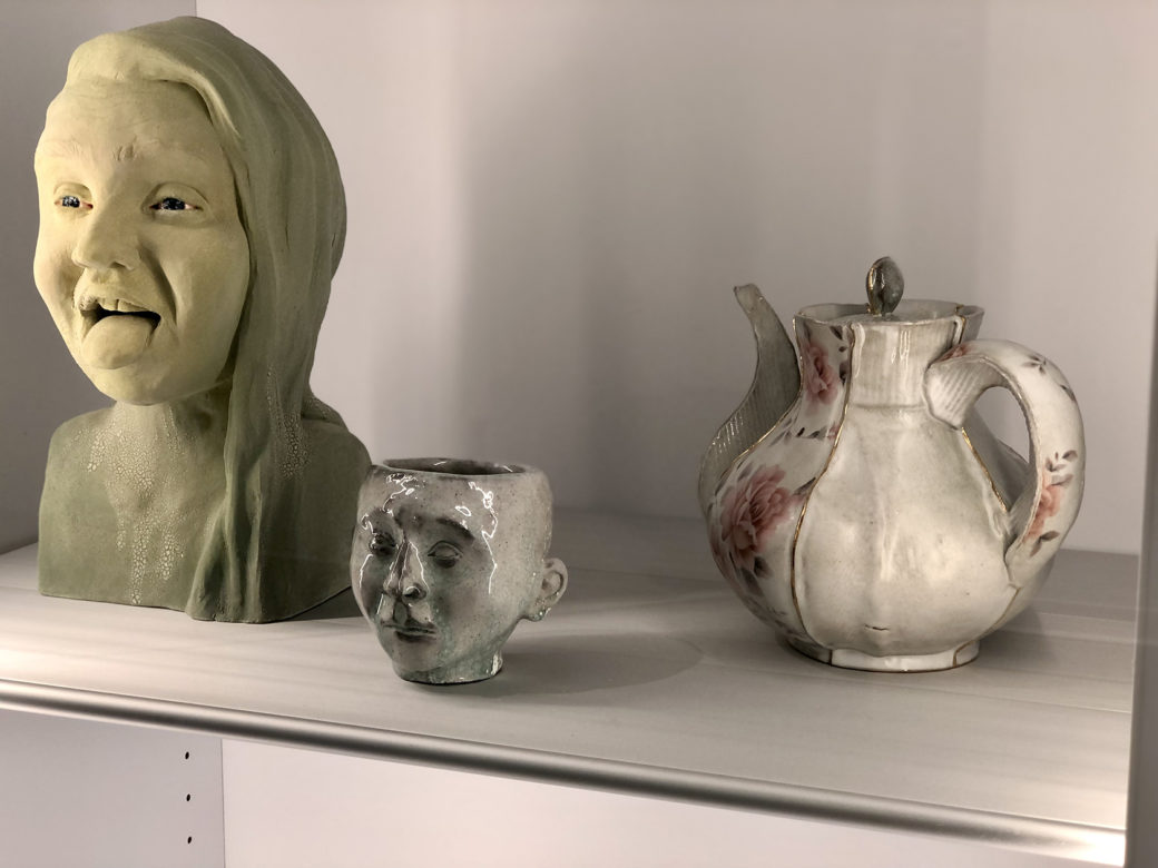 Ceramic artworks -- a bust, a small head vessel and a delicate teapot -- sit on a white shelf