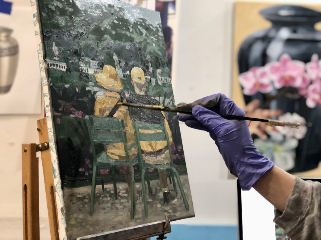 A gloved hand uses a brush to paint an image of a couple on a bench. A drawing and a painting of different vases are visible in the background.