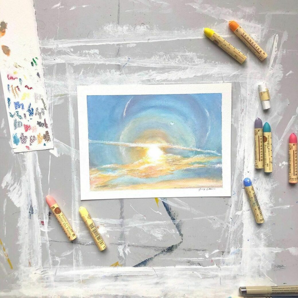 There is an oil pastel drawing of the horizon with a bright blue sky, shining sun and hazy orange and yellow clouds. Blue, yellow, orange, pink and white oil pastels are scattered around the peice.