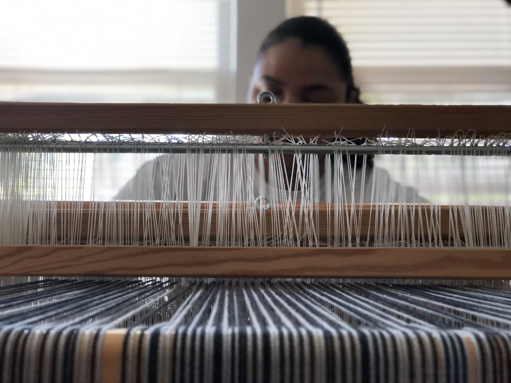 We can see the back of a wooden loom with blue and gray threads held in place. Looking through the loom and slightly out of focus in front of bright windows is the artist. Hair is pulled back and face is turned down looking at the current work in progress