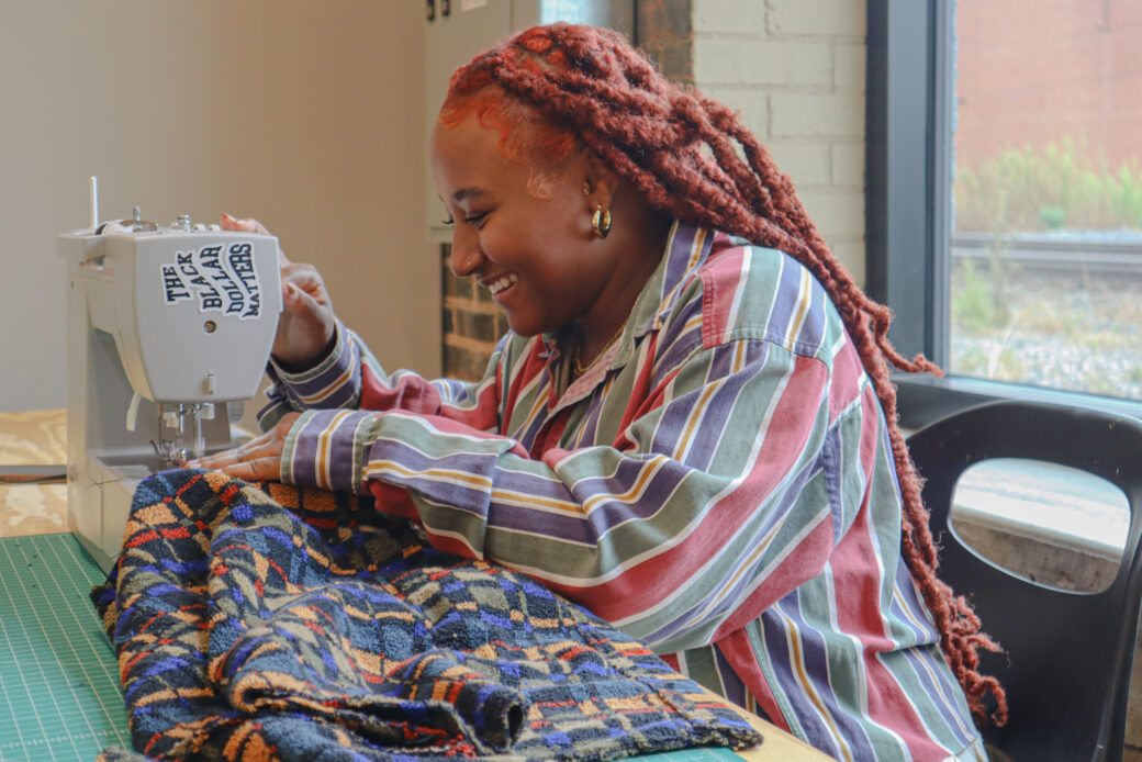 A person with long red braids is smiling, leaning over a gray sewing machine. They are in a striped, multi-color button down shirt, sewing a blue, gold and red patterned piece of fabric.