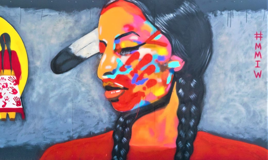 A mural shows a medium-skin toned woman with braids and feathers in her hair and Native American face paint