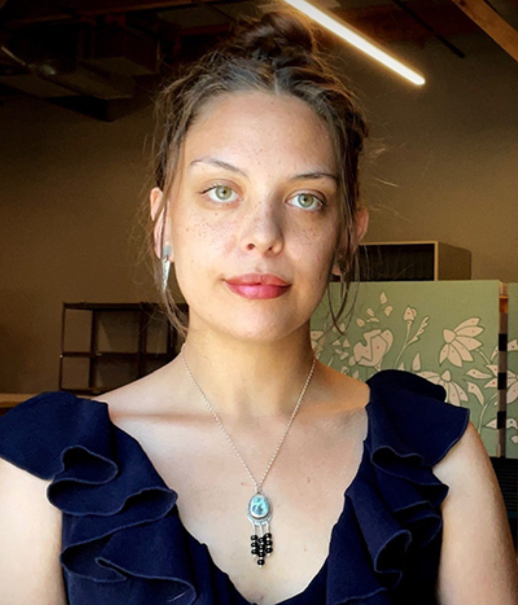 Color photo of a woman wearing a blue shirt and a custom-made jewelry piece smirks at the camera.