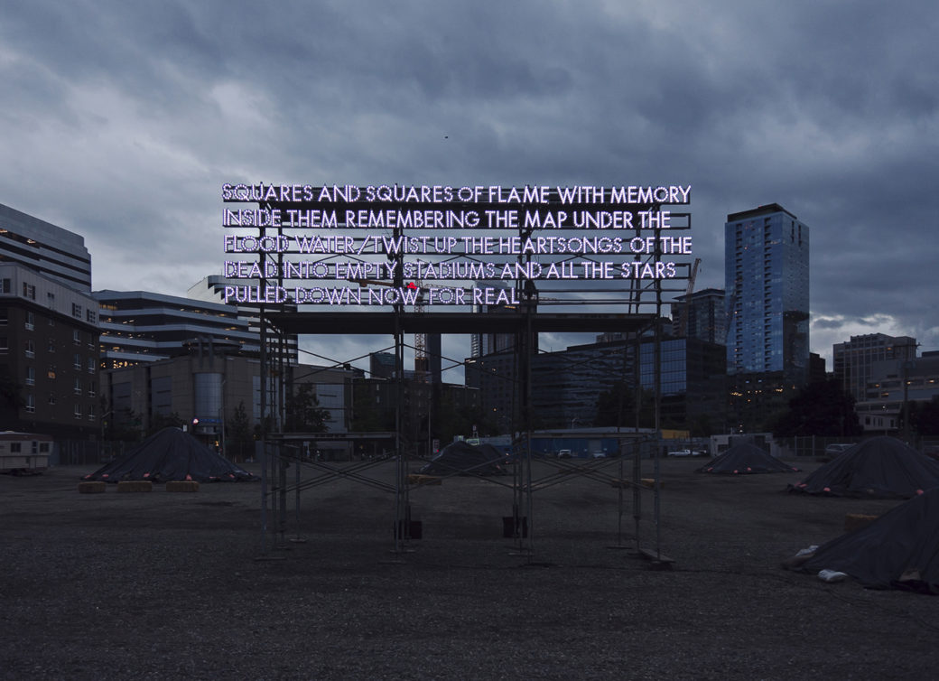 A billboard-size poem in LED light stands on scaffolding in an empty lot, with skyline in the backgroundtands