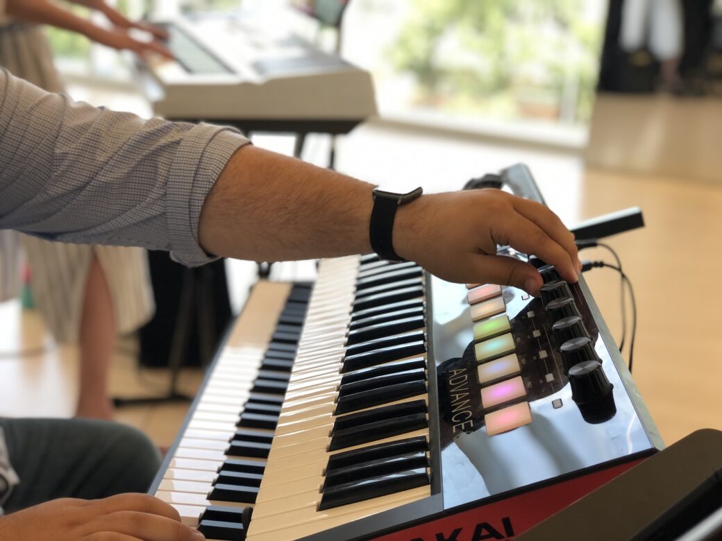 Hands play on a keyboard. The room is bright with natural light.