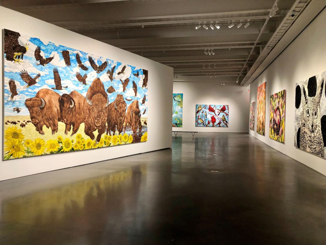 Wide view of seven, large-scale paintings. There are bison charging and eagles soaring against a blue sky, bright red cardinals, two scorpion surrounded by red shapes and yellow lemons and a black and white owl.