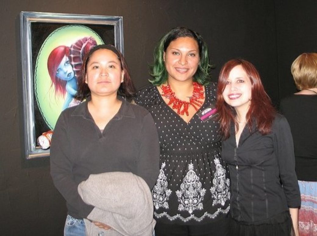 Three younger women stand smiling together, all dressed in black and white. There is an abstract painting hung behind them with bright green and a blue-red figure