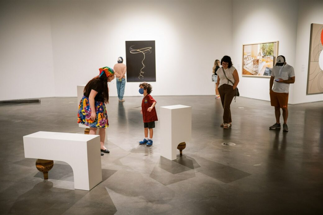 A person is bent over talking to a child. In front of them are two white plinths balancing on ceramic vessels. Behind them is a photo hanging, with a black background and gold lasso in the center.