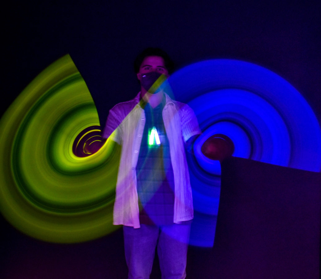 A teen is waving two glow in the dark wands. On the right is blue and on the left is yellow. He is waving them in a circle, and the camera caught the image of incomplete-circle, floating, neon twirls. The rest of the image is dark.