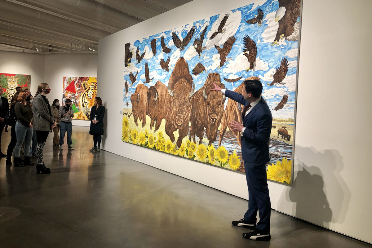 A man in a suit gestures at a large painting of charging bison and swooping eagles while a group of people watches