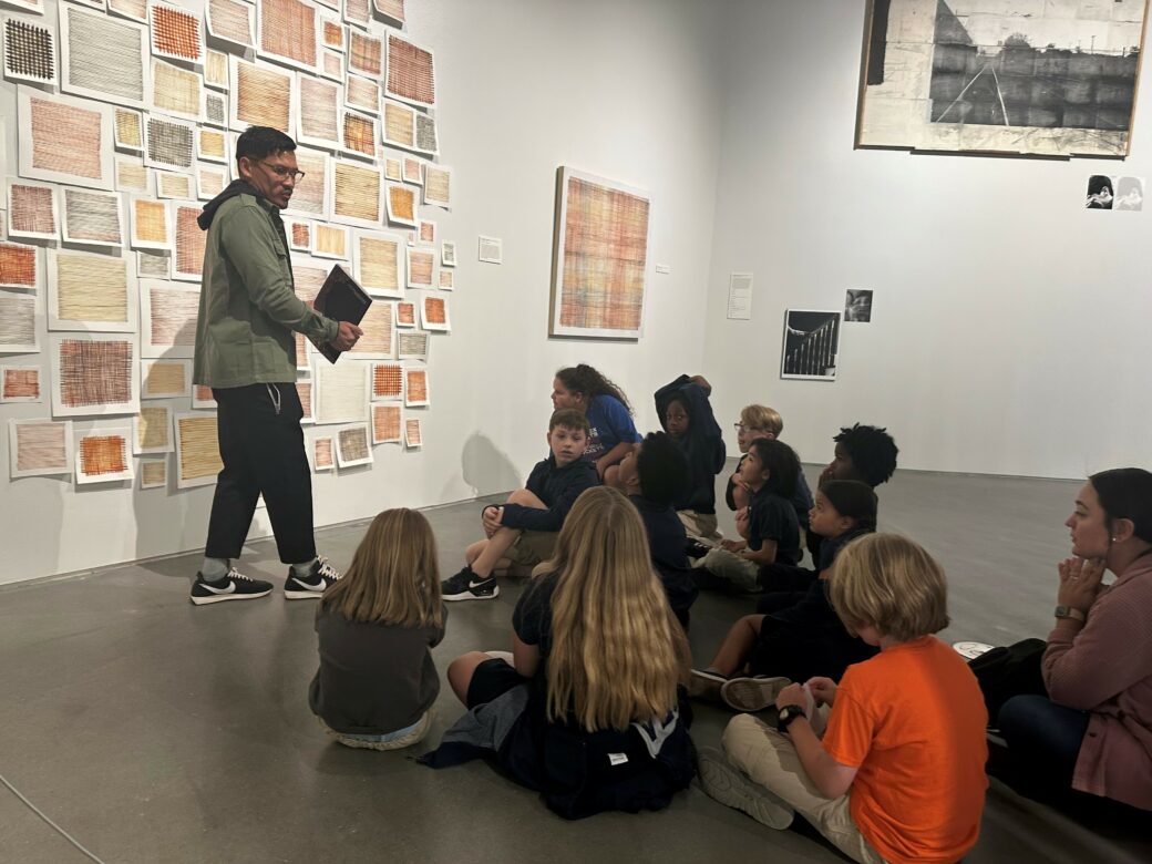 An adult stands in front of a wall of sketches while a group of children sit on the ground in front of them