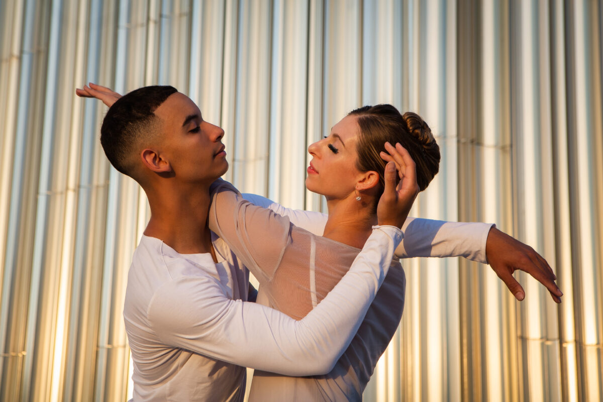 Two dancers stand in a close embrace in front of a silver building