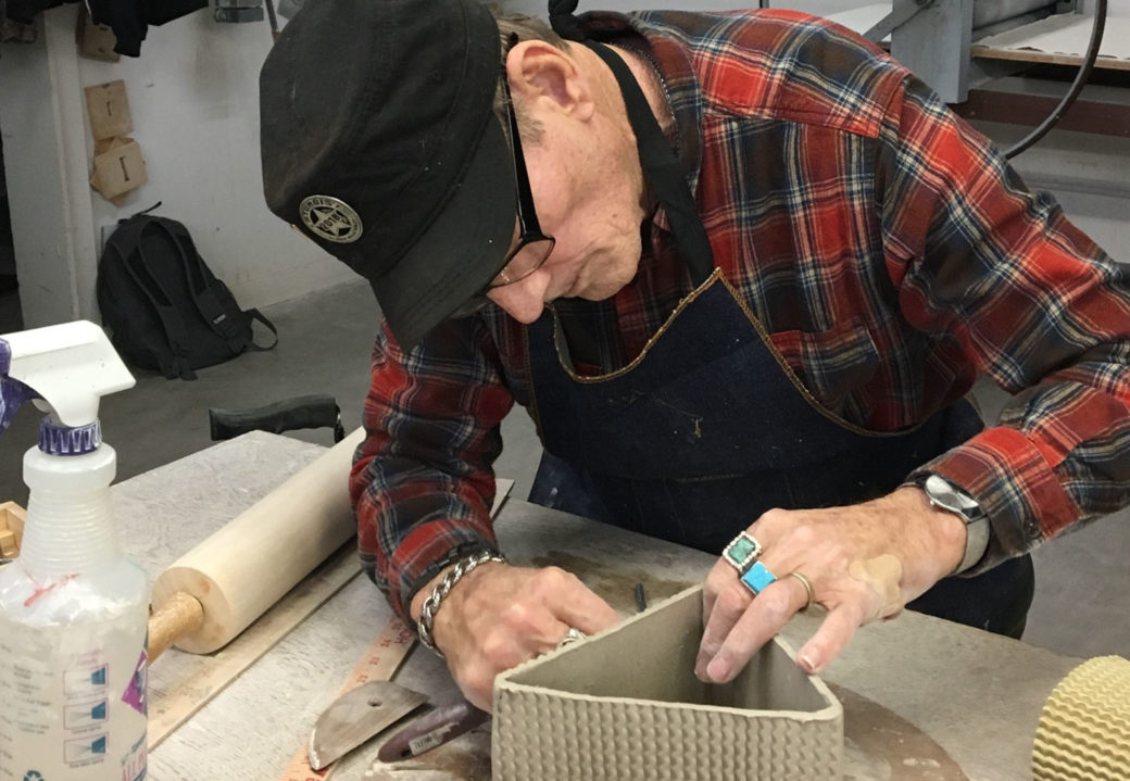 A person in a flannel shirt works on a triangular clay sculpture