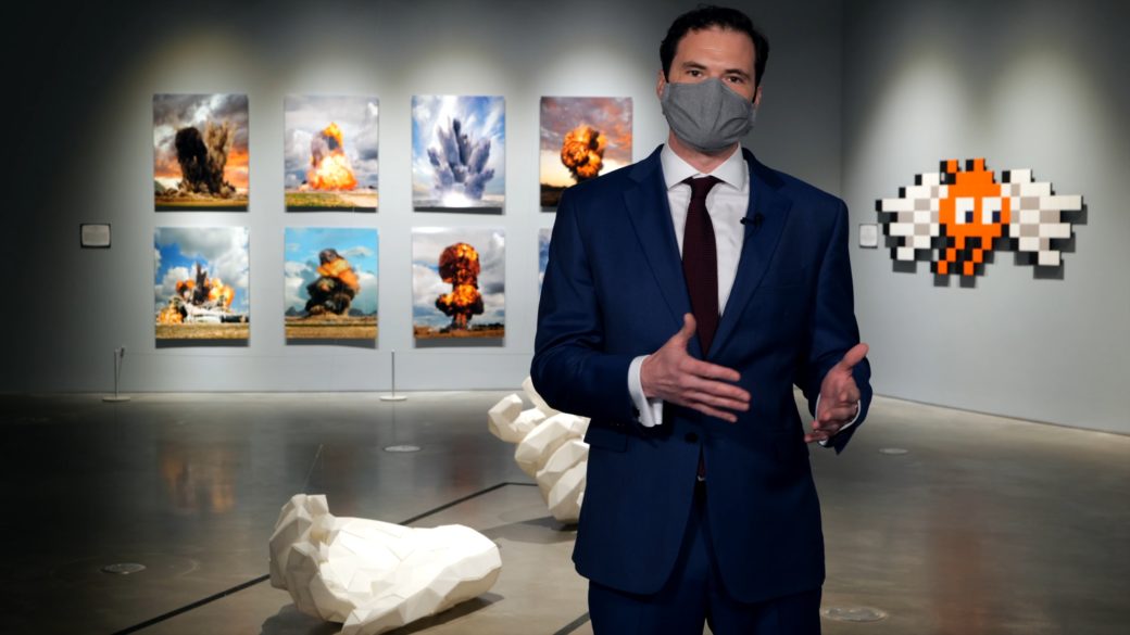 A man in a dark blue suit, tie and mask stands in an art gallery in front of white sculptural hand on the floor and 2-D works on the walls