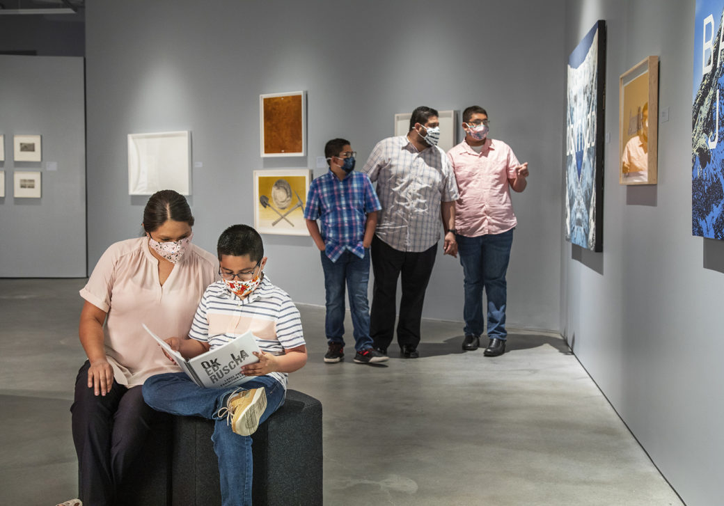 A family in masks in an art gallery -- two figures sit on stools and look at a book, while three others look at art on the walls