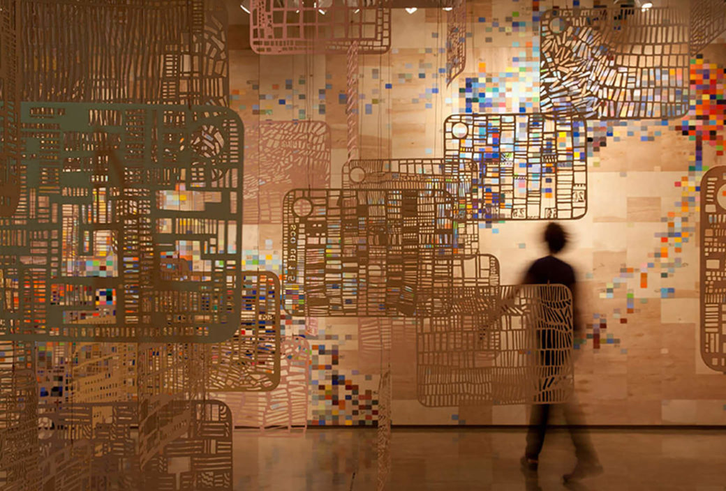A dark figure walks through a gallery filled with flat, suspended sculptures with cutouts in front of a wall covered in pixelated colors