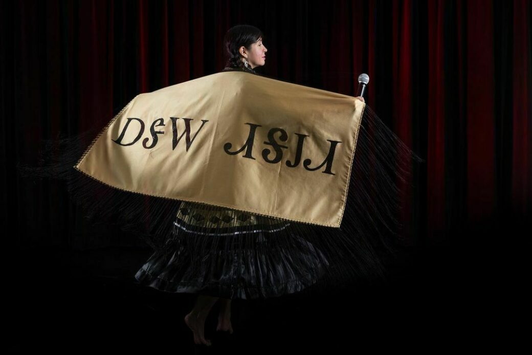 A person with black hair dressed in a dark long skirt against a dark red curtain backdrop is dancing with a gold rectangular cape that has the symbols/word ᎠᏕᎳ ᏗᎦᎫᏗ in large black text.