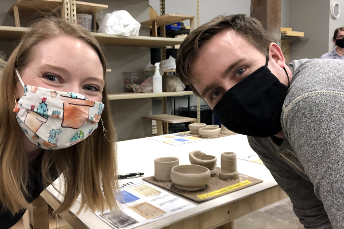A pair of faces in masks look at the camera with pottery in progress behind them
