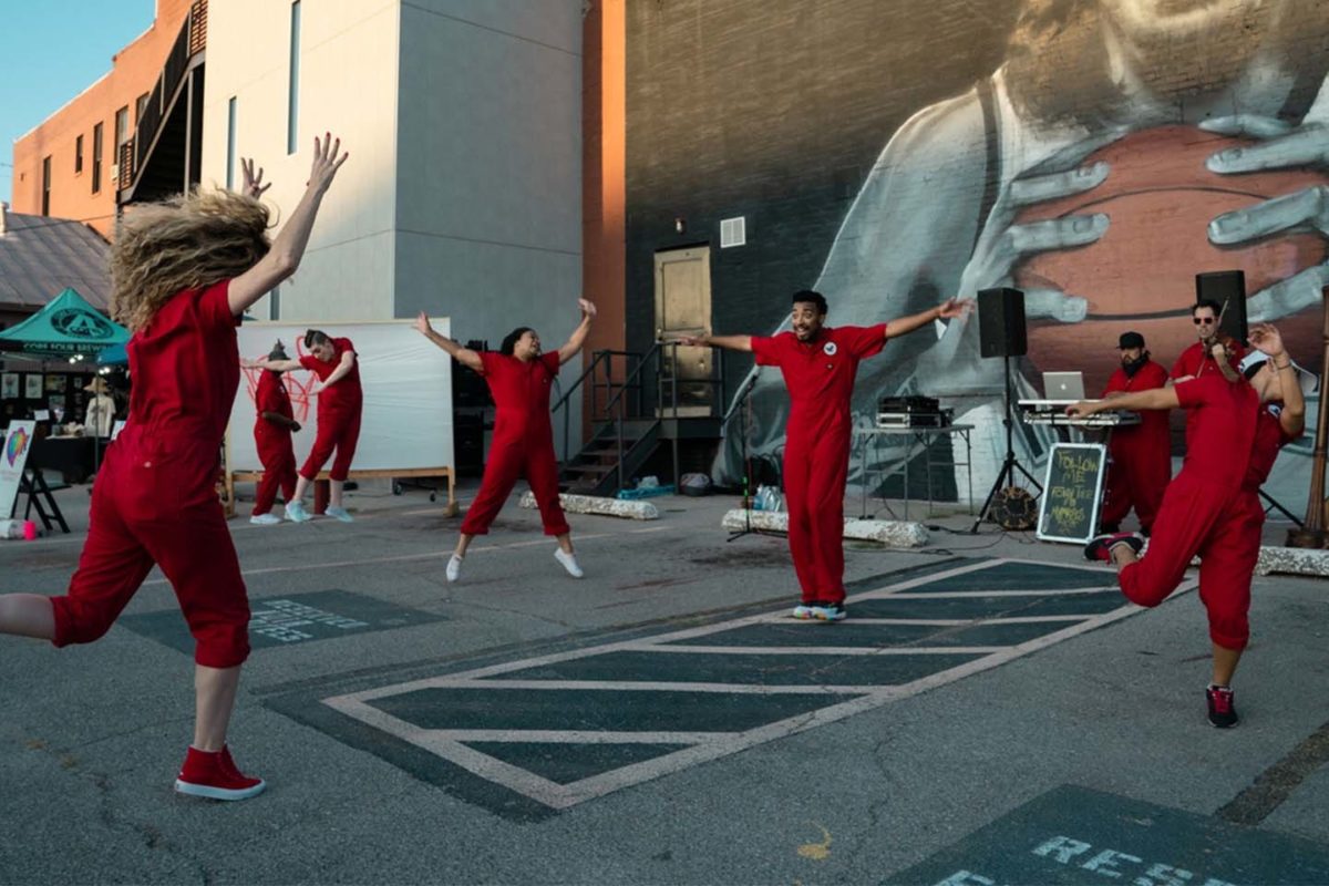 A group of dancers caught mid-movement in matching red jumpsuits in a parking lot overlooked by a large mural of a basketball player