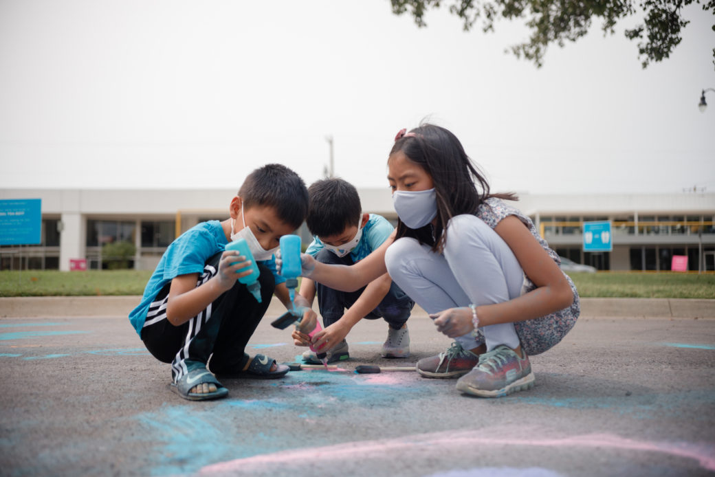 A photo of a group of children in masks drawing with powdered chalk on an asphalt street