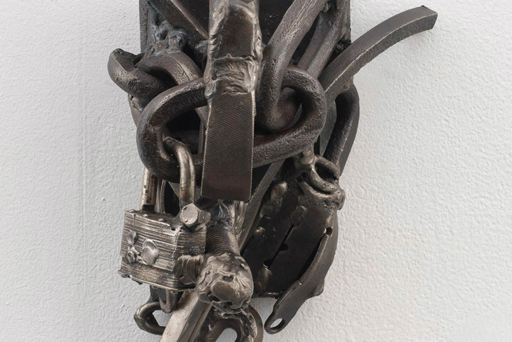 A closeup photo of a sculpture against a white wall with pieces of steel, locks and chain link intertwined