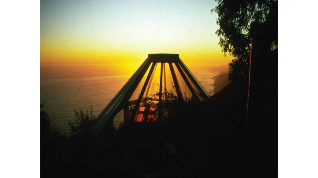 A conical structure made of beams is silhouetted against a sunset