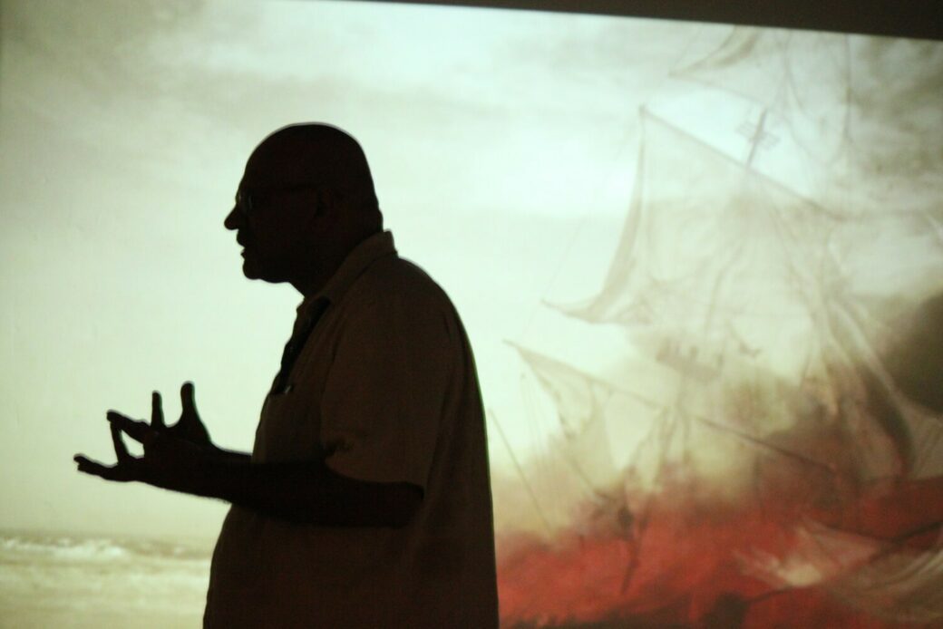 A silhouette of a man pacing and gesturing is visible against a lit backdrop