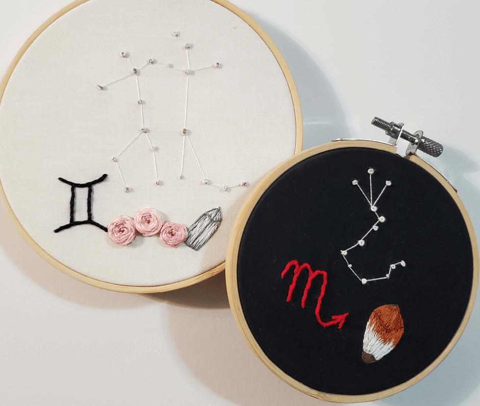 Two embroidery hoops, one with white fabric and Pisces constellation and zodiac symbols, the other with black fabric and Scorpio constellation and symbol.